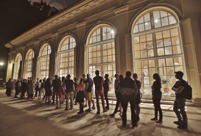 Ludzie stoją wieczorem w kolejce przed oświetloną Starą Oranżerią. 