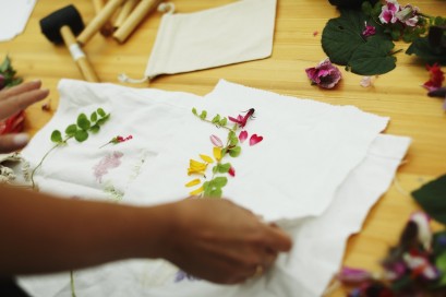 Kobieca dłoń trzyma kawałki białej tkaniny, na której są rozłożone suszone kwiaty. 