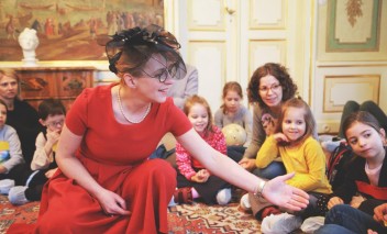 Kobieta w czerwonej sukience, w kapeluszu na głowie, siedzi na podłodze w pokoju, w otoczeniu gromadki dzieci i dwóch kobiet.