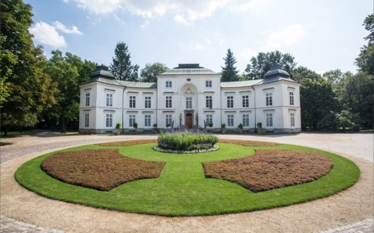 Pałac Myślewicki w Łazienkach Królewskich w otoczeniu drzew. Przed budynkiem znajduje się duży, okrągły trawnik. 