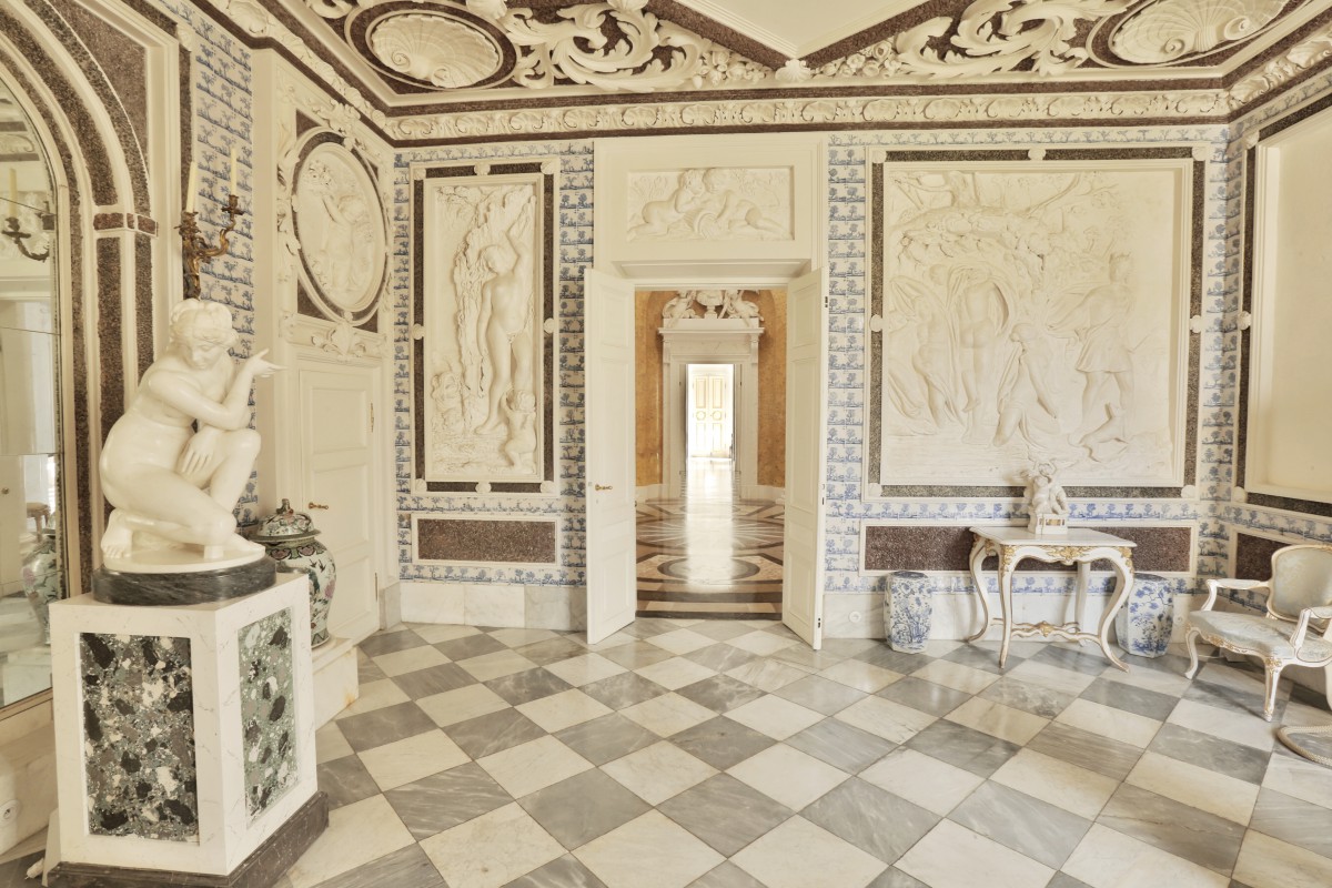 Pokój Kąpielowy w Pałacu na Wyspie. Ściany zdobią płaskorzeźby, pod ścianą po lewej stoi rzeźba przedstawiająca klęczącą kobietę. Pośrodku znajdują się drzwi, po prawej stoją stolik i krzesło.