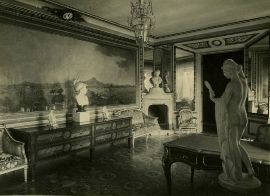 Gabinet Królewski. Po lewej stronie na ścianie wisi obraz i stoi komoda, na której znajduje się rzeźba, dalej jest kominek z rzeźbą, po lewej stronie stoi stolik i posąg nagiej kobiety.
