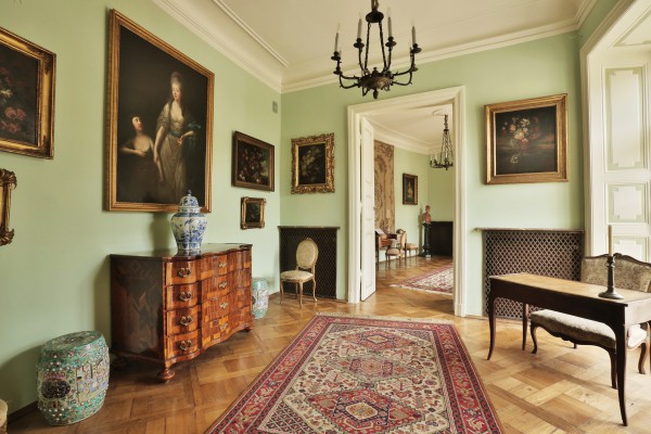Apartament Zachodni w Pałacu Myślewickim, po lewej pod ścianą stoi komoda i wazy, na ścianie wiszą obrazy, pośrodku pomieszczenia znajdują się otwarte drzwi, po pawej stoi stół z krzesłem. 