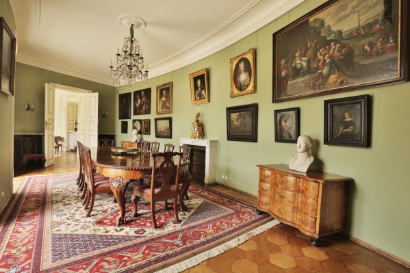 Jadalnia w Pałacu Myślewickim, na środku stoi duży stół z krzesłami, na ścianie po prawej znajduje sie kominek i stoi komoda, ścianę zdobią portrety. 