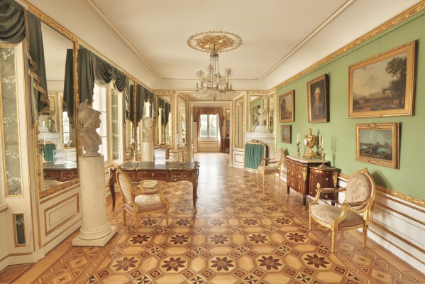 Gabinet Królewski w Pałacu na Wyspie. W pokoju stoi biurko, po lewej stronie są okna, które zdobią upięte kotary, po prawej znajduje się komoda i krzesło, na ścianach wiszą obrazy. 