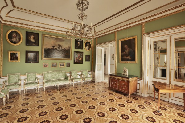 Przedpokój w Pałacu na Wyspie. Na ścianach wiszą portrety, na suficie ozdobny żyrandol, pod ścianą po lewej stronie stoją krzesła, zaś po prawej komoda i stolik.