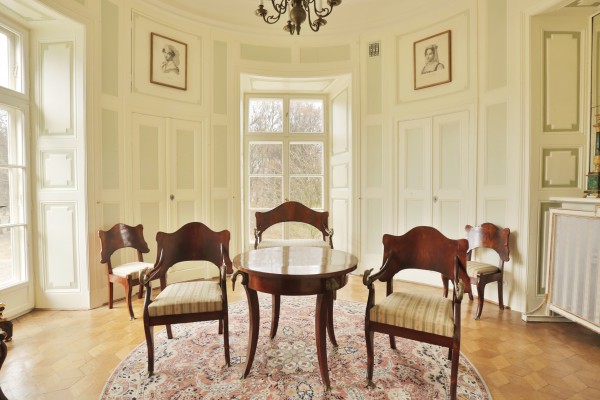 Gabinet narożny w Pałacu Myślewickim, na środku stoi stół z krzesłami, na póokrągłych ścianach znajdują się okna i wiszą obrazki.