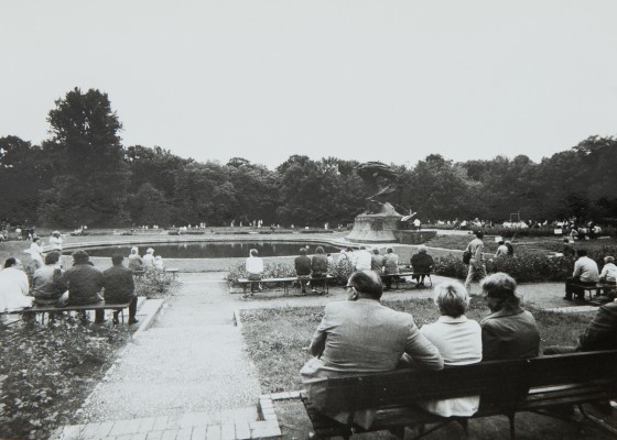 Ludzie siedzą na ławkach przed Pomnikiem Chopina i słuchają koncertu.