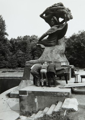 Pomnik Chopina, pod monumentem stoi trzech mężczyzn odwróconych tyłem, którzy stawiają fortepian.