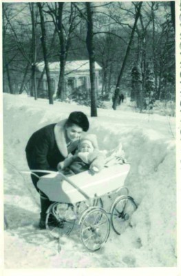 Zima w parku, kobieta nachyla się nad wózkiem i podnosi dziecko. 