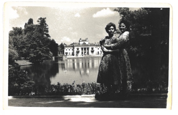 Dwie kobiety pozują do zdjęcia w łazienkowskich ogrodach, w tle widać Pałac na Wyspie w otoczeniu wody i drzew.