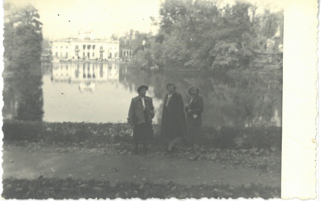 Trzy kobiety stoją na ścieżce w tle widać Pałac na Wyspie w otoczniu wody i drzew. 