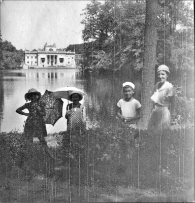 Dwie dziewczynki z parasolami w rękach, chłopiec i kobieta stoją na parkowej ścieżce, przy stawie, w tle widać Pałac na Wyspie.