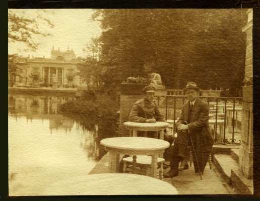 Archiwalne zdjęcie przedstawiające dwóch mężczyzn siedzących przy stoliku na tarasie Amfiteatru w Łazienkach Królewskich, za nimi rozciąga się woda, w tle Pałac na Wyspie w otoczeniu zieleni.