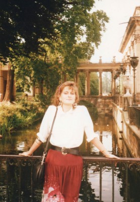 Kobieta ubrana w białą bluzkę i czerwoną spódnicę stoi przodem, oparta o poręcz mostka nad kanałem przy Pałacu na Wyspie.