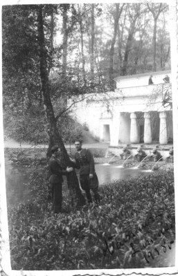 Dwóch mężczyzn w mundurach wojskowych stoi w parku pod drzewem. Opierają się rękami o pień. W tle widoczne są kolumny Świątyni Egipskiej w Łazienkach Królewskich.