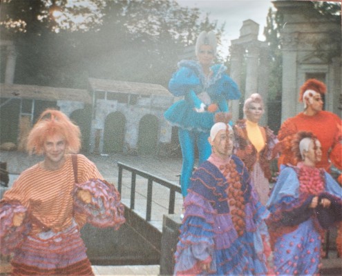 Aktorzy teatralni w kolorowych strojach i perukach na głowie stoją w Amfiteatrze w Łazienkach Królewskich.