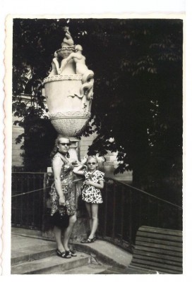 Kobieta i dziewczynka stoją na schodach przy poręczy, w tle rzeźbiona latarnia i drzewa. 