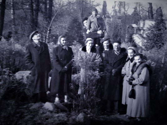Archiwalne zdjęcie przedstawiające grupę kobiet i mężczyzn w zimowych płaszczach, którzy stoją w ogrodzie Łazienek Królewskich. Mężczyzna stojący z tyłu unosi do góry chłopca.