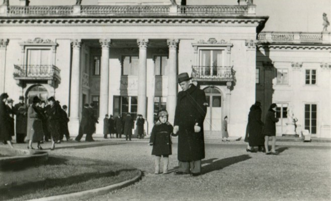 Kilkuletni chłopiec i mężczyzna stoją przed Pałacem na Wyspie w Łazienkach Królewskich. Mają na sobie zimowe płaszcze, mężczyzna nosi kapelusz. Przed Pałacem na Wyspie spacerują ludzie.