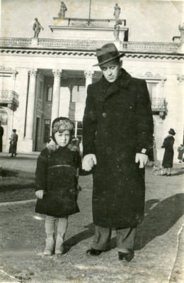  Kilkuletni chłopiec i mężczyzna stoją przed Pałacem na Wyspie w Łazienkach Królewskich. Mają na sobie zimowe płaszcze, mężczyzna nosi kapelusz. Przed Pałacem na Wyspie spacerują ludzie.