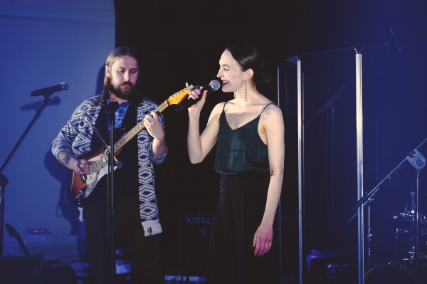 Kobieta stoi na scenie przy mikrofonie i śpiewa, obok stoi mężczyzna grający na gitarze. 