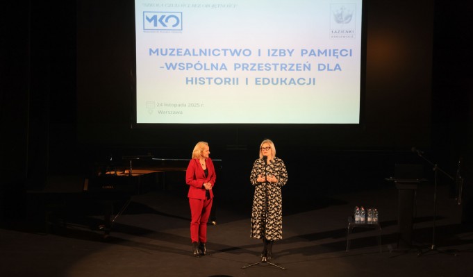 Dwie kobiety stoją na scenie. Kobieta po lewej jest ubrana w czerwone spodnie i marynarkę, a kobieta po prawej w czarną sukienkę w białe wzory. Powyżej, nad ich głowami, wisi plansza z napisem "Muzealnictwo i izby pamięci - wspólna przestrzeń dla historii"