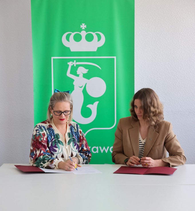 Dwie kobiety siedzą przy stole, mają przed sobą rozłożone kartki. Kobieta po lewej podpisuje jedną z kartek. 