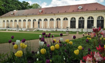 Budynek Starej Oranżerii w Łazienkach Królewskch, widać fragment ogrodu, w którym kwitną żółte, białe i bordowe kwiaty.