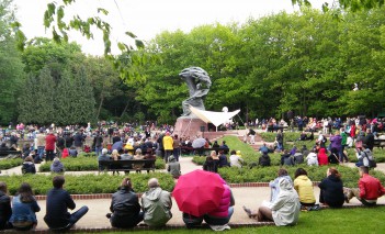 Ludzie siedzą na trawie wokół Pomnika Chopina i słuchają koncertu.
