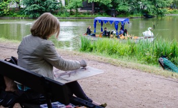 Kobieta siedzi w parku i maluje, przed nią znajduje się rzeka, po której płynie gondola.