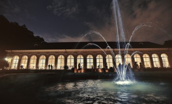 Stara Oranżeria nocą, oświetlona wewnątrz światłem, z przodu budynku widoczna jest tryskająca fontanna.