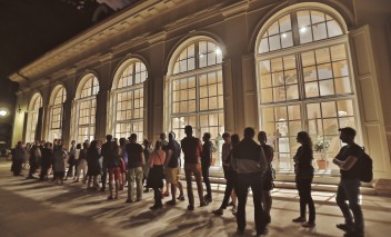 Ludzie stoją wieczorem w kolejce przed oświetloną Starą Oranżerią. 