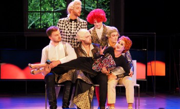 Aktorzy na scenie w kolorowych perukach, trzech z nich siedzi, trzymając leżącą im na kolanach kobietę, dwóch stoi z tyłu.