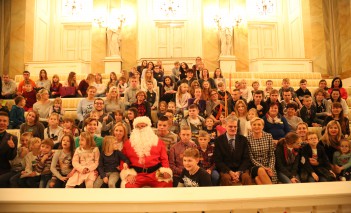 Grupa kilkudziesięciu dorosłych i dzieci siedzących na widowni Teatru Królewskiego, pośrodku nich siedzi święty Mikołaj. 