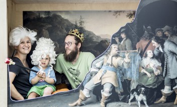 Kobieta w peruce, dziecko w peruce i mężczyzna w koronie na tle obrazu pozują do zdjęcia.