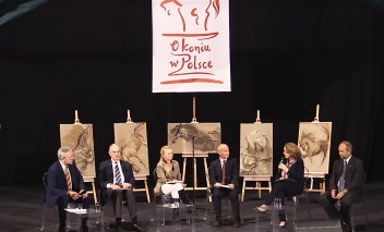 Grupa pięciu osób siedzi na krzesłach ustawionych na scenie i rozmawia, w tle za nimi ustawione są na sztalugach obrazy koni.
