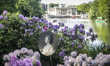Widok na Pałac na Wyspie wśród wody i zieleni. Na pierwszym planie pośród fioletowych kwiatów wetknięta została reprodukacja portretu z kolekcji Stanisława Augusta. 