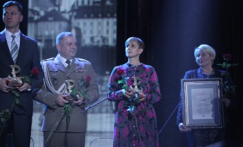 Na scenie, na tle zdjęcia fragmentu miasta, stoją dwaj mężczyźni i dwie kobiety, trzymają statuetki i róże, a ostatnia z kobiet - dyplom.