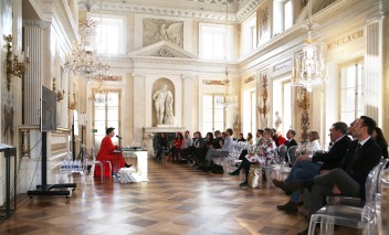 Konferencja w Pałacu na Wyspie. Uczestnicy siedzą na krzesłach, na przeciwko nich siedzi prelegent - kobieta ubrana w długą czerwoną sukienkę.