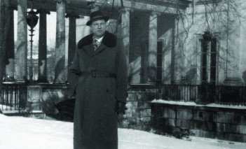 Mężczyzna w płaszczu i kapeluszu stoi przed Pałacem na Wyspie zimą. 