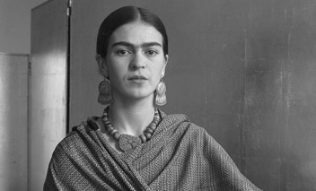 Czarno-białe, archiwalne zdjęcie Fridy Kahlo. Artystka ubrana jest w sukienkę i chustę, którą ma owinięte ramiona, oparta jest o kanapę, prawy łokieć trzyma na wiklinowym, plecionym koszyku. 