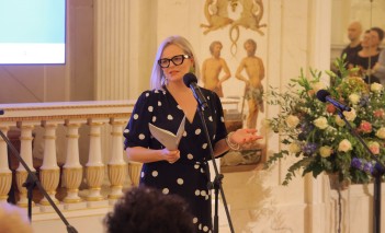 Kobieta w sukience stoi przed mikrofonem i przemawia. 