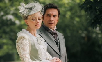 Kobieta w długiej, białej, koronkowej sukni i kapeluszu spaceruje obok mężczyzny w garniturze na tle parku.