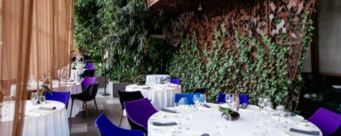 Wnętrze restauracji, w której stoją okrągłe stoły nakryte białymi obrusami, wokół nich ustawione zostały niebieskie krzesła.