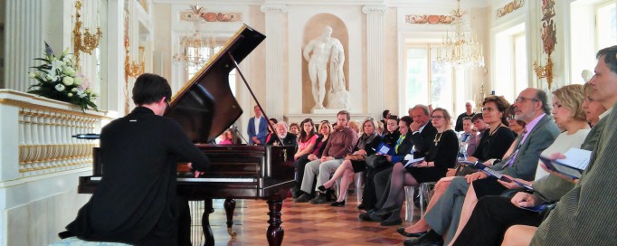 Mężczyzna gra na fortepianie, ludzie siedzą na krzesłach i słuchają koncertu.