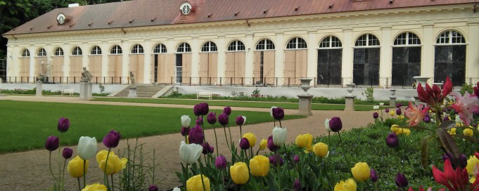 Budynek Starej Oranżerii w Łazienkach Królewskch, widać fragment ogrodu, w którym kwitną żółte, białe i bordowe kwiaty.