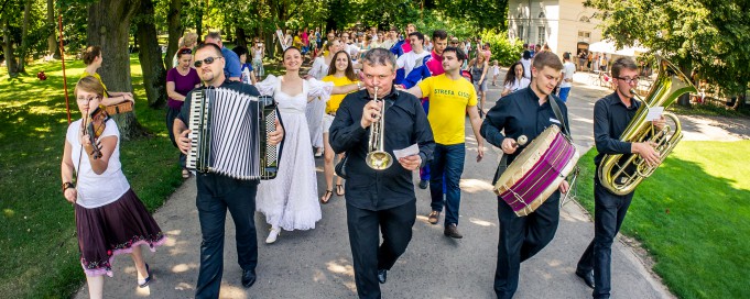 Grupa muzyków gra na instrumentach, idąc parkową aleją.