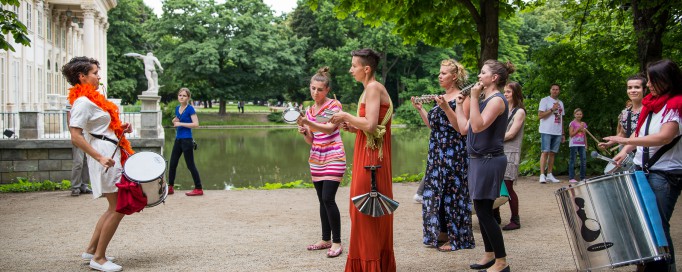 Kobieta gra na bębnie, dwie inne stoją przed nią i klaszczą.