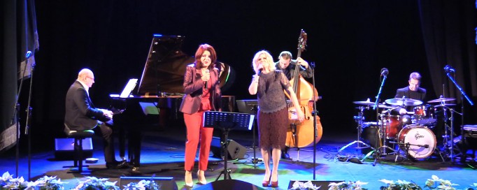 Dwie kobiety stoją na scenie przed mikrofonem i śpiewają, jeden mężczyzna gra na fortepianie, drugi na wiolonczeli.